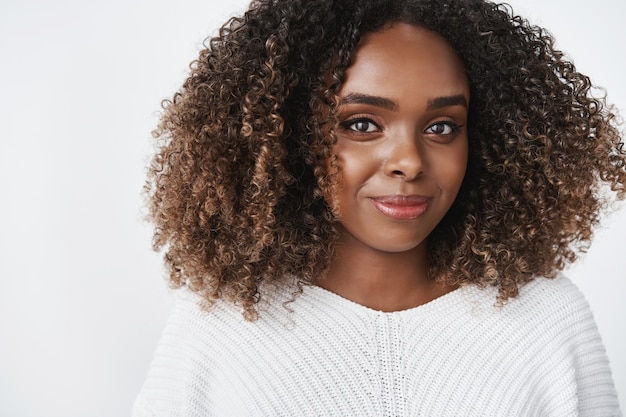 Close-up de uma jovem afro-americana de 25 anos, satisfeita, relaxada e alegre, carismática, com cabelo encaracolado, sorrindo com sinceridade e olhando para a frente despreocupada e encantada com um olhar terno