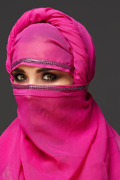 Close-up de uma jovem bonita com olhos esfumaçados expressivos usando o hijab rosa chique decorado com lantejoulas. Ela virou a cabeça e olhando para a câmera em um fundo escuro. Hu