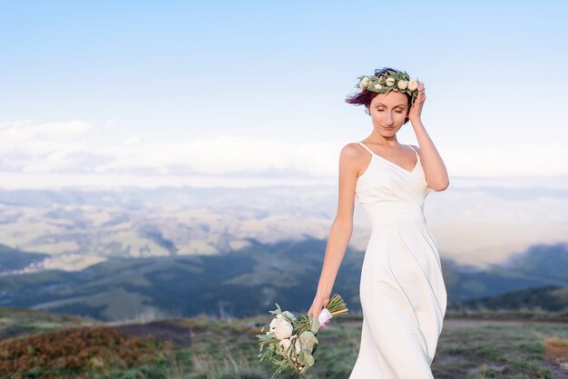 Close-up de uma jovem em um vestido branco com uma coroa de flores na cabeça e um buquê de flores ao ar livre