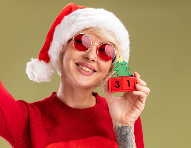 Close-up de uma jovem loira sorridente usando chapéu de natal e suéter de natal de papai noel com óculos segurando um brinquedo de árvore de natal com data parecendo isolado na parede verde oliva