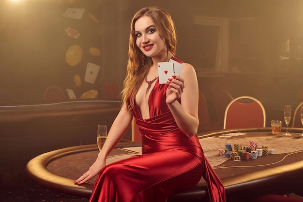 Close-up de uma loira maravilhosa em um vestido longo de cetim vermelho, com dois ases na mão posando sentado em uma mesa de pôquer no cassino de luxo. Paixão, cartas, fichas, álcool, vitórias, jogos de azar - é af