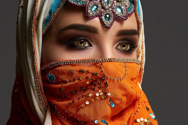 Close-up de uma menina bonita com maquiagem profissional usando um hijab colorido decorado com lantejoulas e joias. Ela está posando no estúdio e olhando para longe em um fundo escuro. Emoções humanas,