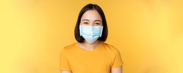 Close-up de uma mulher asiática feliz sorridente usando máscara facial médica da posição covid em camiseta amarela