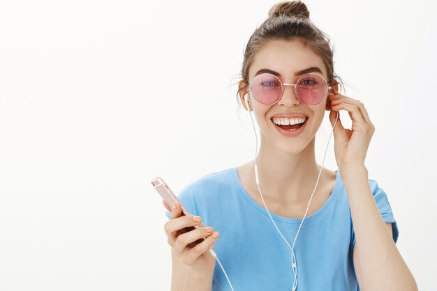 Close-up de uma mulher atraente e moderna em óculos de sol, ouvindo música, curtindo a música nos fones de ouvido, segurando o smartphone