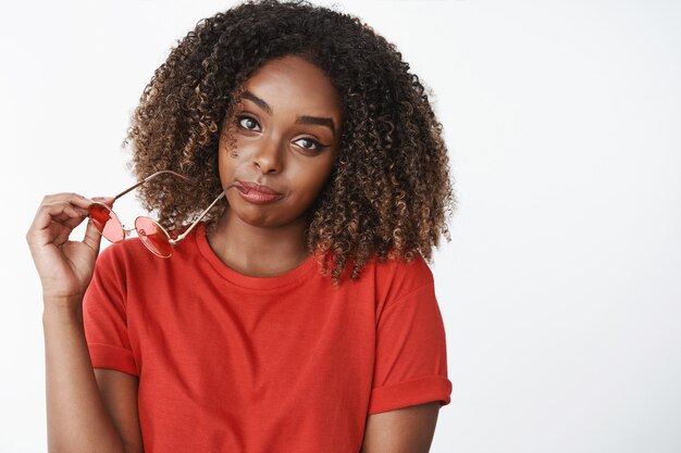 Close-up de uma mulher esperando pelo namorado inclinando a cabeça, mordendo a borda dos óculos e parecendo boba e casual na frente usando uma camiseta vermelha brilhante posando duvidosa com as sobrancelhas levantadas sobre a parede branca