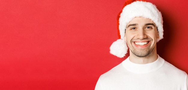Close-up do homem sorridente atraente no suéter branco e chapéu de Papai Noel, parecendo feliz aproveitando o inverno holid.
