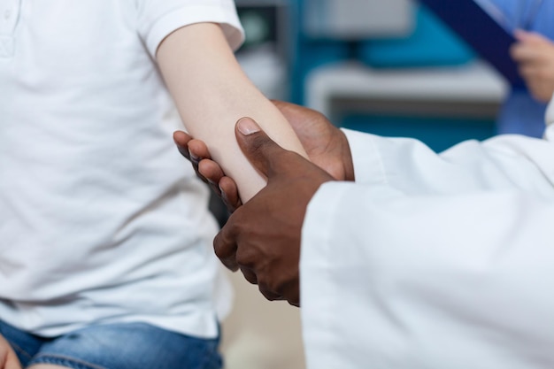 Close-up do médico pediatra americano africano esfregando o braço ferido do pequeno paciente durante a fisioterapia médica no escritório do hospital. Criança com osso quebrado após acidente. Serviço de saúde