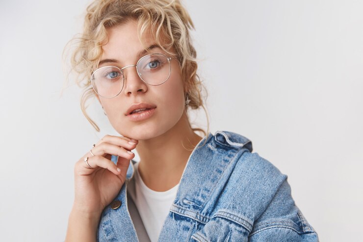 Foto grátis close-up, elegante, bonita, elegante, loira, olhos azuis, blogueira, óculos transparentes, jaqueta jeans, tocando o queixo, câmera pensativa, intrigada, interessada, parede branca