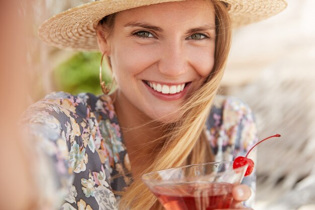 Close-up foto de bela jovem tem festa de verão com amigos, gosta de beber uma bebida saborosa de frutas, faz selfie com um sorriso alegre, vestido com roupas da moda