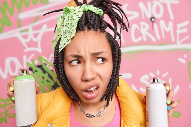 Close-up, foto de indignada insatisfeita garota adolescente afro-americana com lábios concentrados ao lado, penteado com dreadlock segura garrafas de aerossol, pinturas criativas na parede da rua