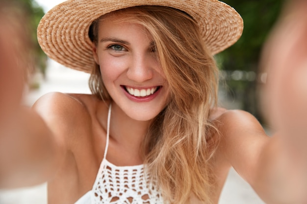 Close-up foto de uma bela jovem satisfeita fazendo selfie, tem uma expressão feliz, usa chapéu de palha e vestido branco de verão, feliz por posar para a câmera e se fotografar, expressa positividade