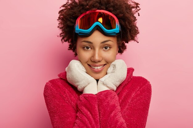 Close-up foto de uma jovem bonita com máscara de snowboard, mantém as duas mãos sob o queixo, usa luvas brancas quentes, olha diretamente para a câmera.