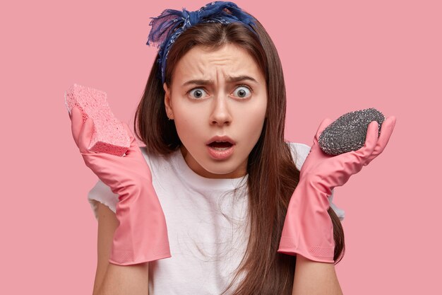 Close-up foto de uma jovem perplexa com expressão facial surpresa, carregando esponja, fazendo tarefas domésticas, maravilhada com a quantidade de trabalho