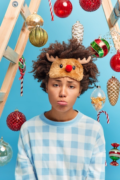 Foto grátis close-up foto de uma mulher afro-americana descontente com uma expressão sombria no rosto para a câmera triste por estar doente durante as férias de inverno usa uma máscara de dormir quente e um pijama decora a casa com árvores de ano novo