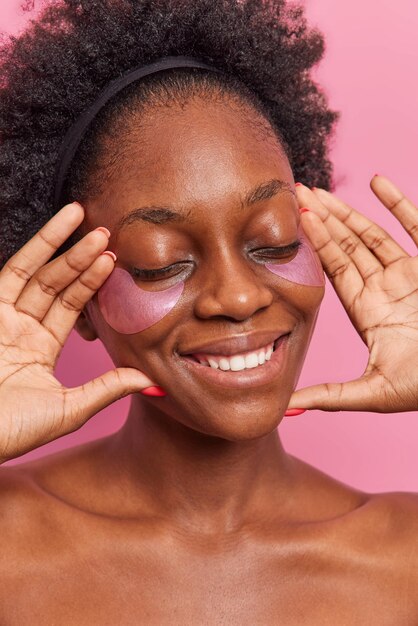 Close-up foto de uma mulher gentil satisfeita com pele escura aplicando adesivos de hidrogel sob os olhos ansiosos para ter uma pele facial fresca e ombros nus fica feliz por tentar reduzir as bolsas nos olhos. Conceito de beleza