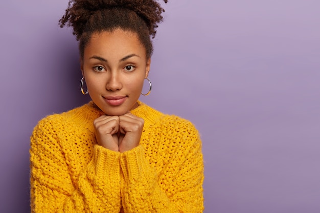 Close-up foto de uma mulher séria e bonita de cabelo encaracolado focalizou a expressão para a câmera, aparência confiante usa um suéter amarelo com as duas mãos embaixo do queixo