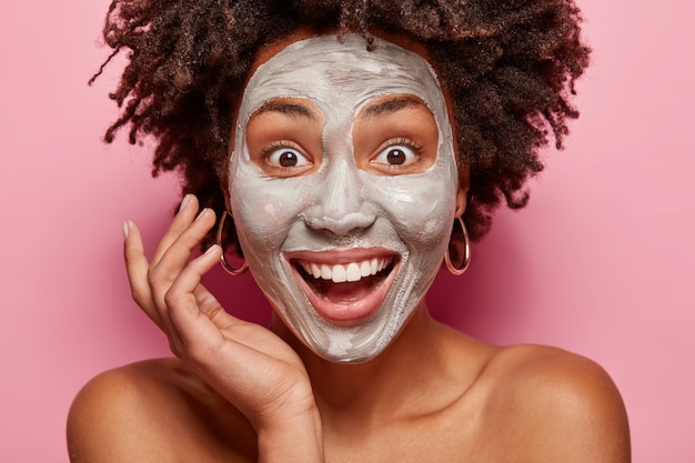 Close up retrato da encantada senhora afro-americana com máscara de argila branca no rosto, sorri amplamente, surpresa por ter a pele fresca após procedimentos de beleza, tem consulta com esteticista ou cosmetologista