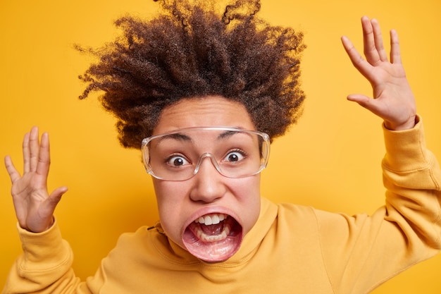 Close-up retrato de uma mulher de cabelo encaracolado emocional olha fixamente, tem uma boca muito grande, levanta os braços, usa óculos grandes de óptica