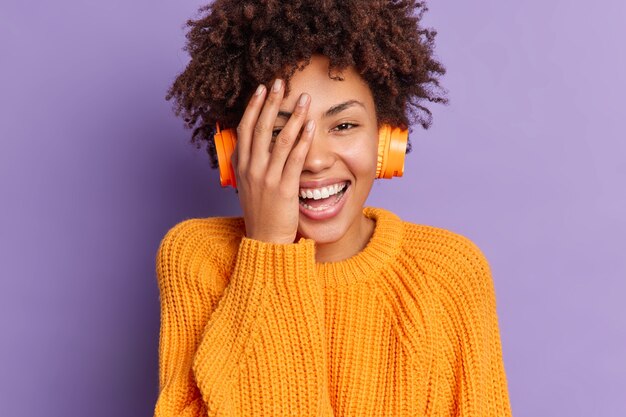 Close-up retrato de uma mulher de pele escura radiante mantém a mão no rosto e sorri despreocupada ouve música favorita em fones de ouvido vestida casualmente expressa emoções positivas