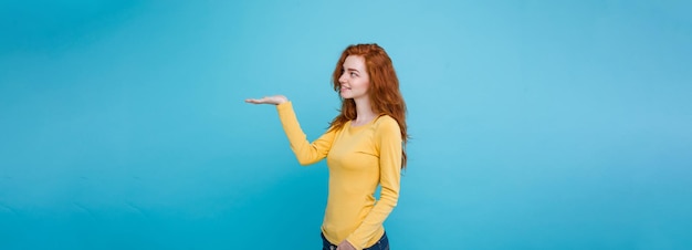 Foto grátis close-up retrato jovem linda garota ruiva atraente feliz com algo e apontando o dedo fundo pastel azul copiar espaço