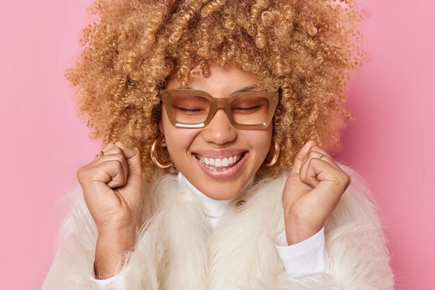 Close-up tiro de alegre jovem bonita com cabelo encaracolado mantém os olhos fechados sorrisos amplamente cerra os punhos celebra o sucesso usa óculos e casaco de pele branco isolado sobre fundo rosa