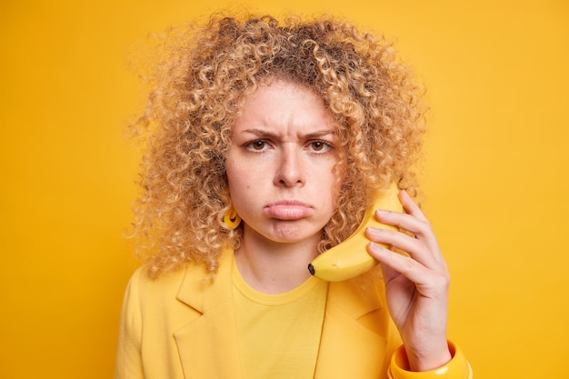 Close-up tiro de mulher de cabelo encaracolado taciturno segura banana enquanto o telefone perto da orelha finge que ligar para alguém tem expressão descontente vestida com roupas amarelas expressa emoções negativas poses interiores.