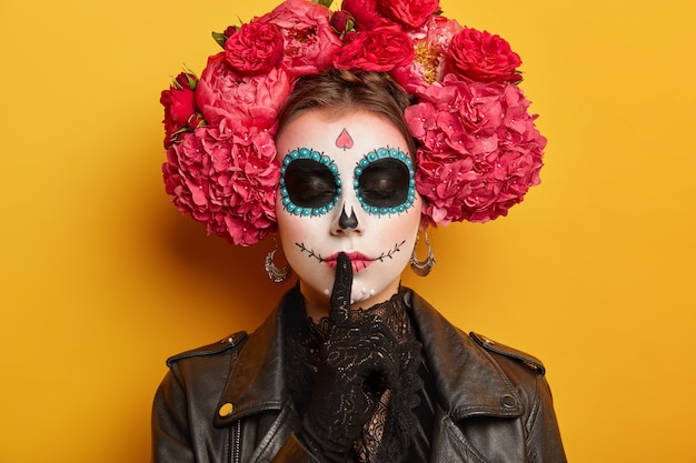 Close-up tiro de mulher secreta tem açúcar caveira maquiagem faz gesto de silêncio mantém o dedo sobre os lábios, fica com os olhos fechados pintados em círculos escuros em torno de ser charmoso e perigoso.