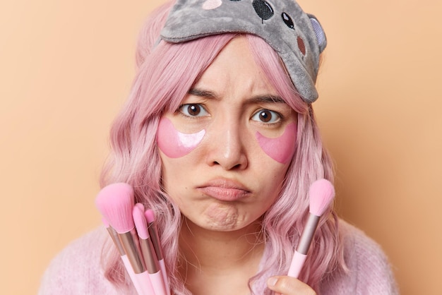 Foto grátis close-up tiro de triste mulher asiática descontente franze os lábios parece ofendido com a câmera usa pincéis cosméticos para aplicar maquiagem coloca em manchas de hidrogel sob os olhos usa máscara de dormir na testa.