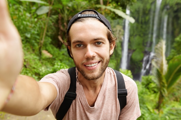 Close-up vista de um alpinista caucasiano bonito usando snapback olhando com um sorriso feliz enquanto tira uma selfie com uma paisagem incrível com cachoeira