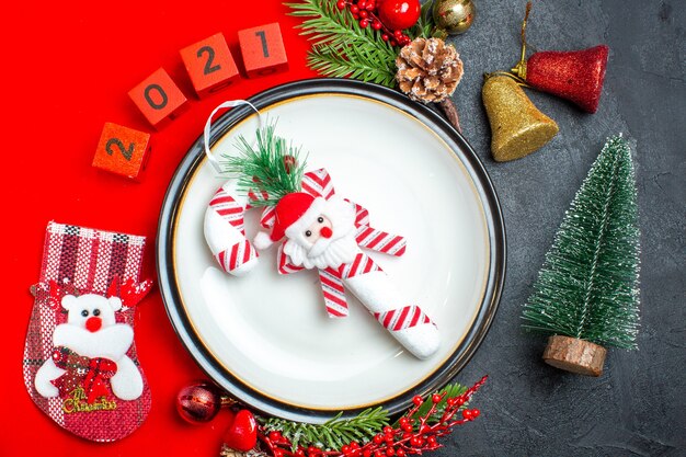 Close-up vista do fundo de ano novo com acessórios de decoração de prato de jantar ramos de abeto e números meia de Natal em um guardanapo vermelho ao lado da árvore de Natal em uma mesa preta