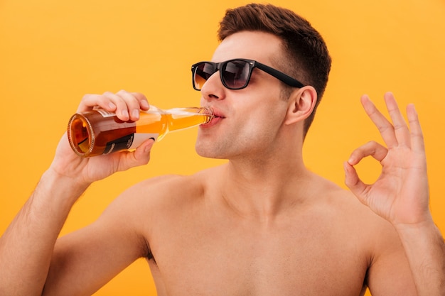 Close-up vista do homem nu sorridente em óculos de sol, bebendo cerveja e mostrando sinal bem amarelo