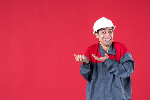 Close-up vista do jovem arquiteto sorridente de uniforme com capacete na parede vermelha isolada