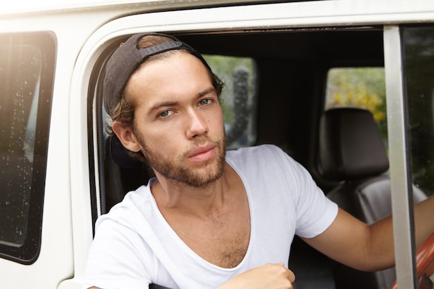 Foto grátis close-up vista do jovem bonito com barba elegante, sentado no banco do motorista na cabine de couro de seu carro branco com tração nas quatro rodas e olhando com expressão séria durante a viagem