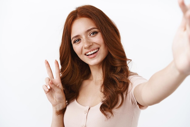 Closeup amigável ruiva otimista com sardas na blusa segure a câmera uma mão tomando selfie no smartphone parecendo positivo e alegre mostrar paz bem-estar sinal de vitória fotografando