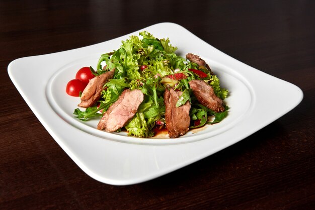 Closeup com fome de um bife grelhado fatiado servido com salada de folhas verdes e tomate cereja em um prato quadrado branco em cima da mesa no restaurante.