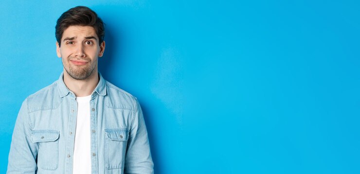 Foto grátis closeup de cara cético e desajeitado sorrindo maliciosamente sentindo-se desconfortável em pé sobre fundo azul