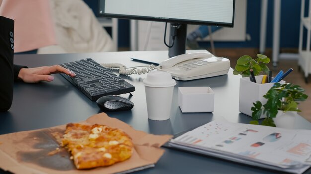 Closeup de empresária sentada na mesa em frente ao computador comendo uma fatia de pizza enquanto fala no telefone fixo com o gerente remoto da empresa