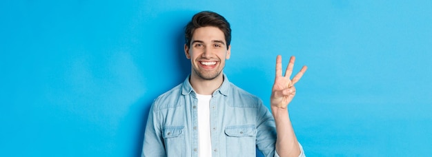 Closeup de homem bonito sorrindo mostrando os dedos número três em pé sobre fundo azul