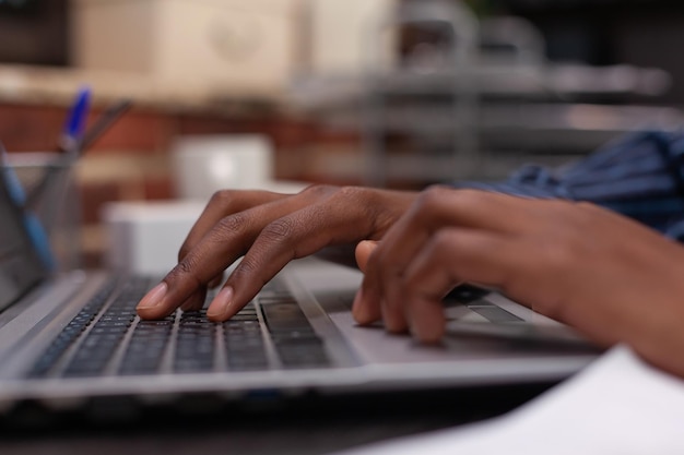 Closeup de mãos afro-americanas digitando dados de negócios no teclado de computador portátil no escritório de parede de tijolos de inicialização. Concentre-se no funcionário de inicialização usando laptop para trabalho profissional ou redigindo e-mail.