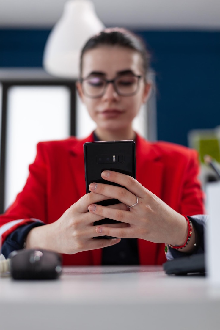 Foto grátis closeup de mãos com smartphone sobre mesa de escritório de inicialização. mulher de negócios com óculos segurando telefone inteligente em ambiente de negócios. empresário em conteúdo de navegação de jaqueta vermelha no dispositivo móvel.