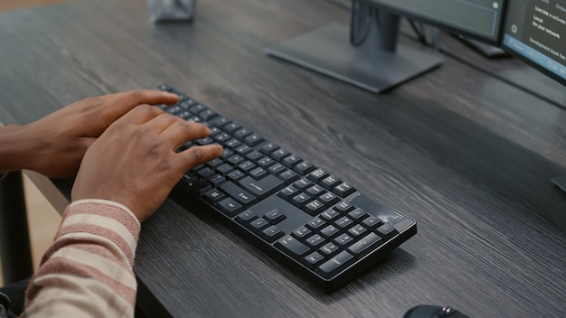 Closeup de mãos de programador americano africano digitando código no teclado enquanto olha para telas de computador com interface de programação. Engenheiro de sistema sentado na mesa desenvolvendo algoritmo.