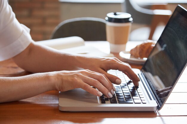 Closeup de mãos femininas, digitando no laptop com copo para viagem e croissant