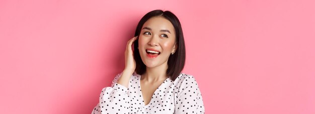 Closeup de mulher asiática glamour suspirando, olhando para a esquerda sonhadora e sorrindo em pé sobre um fundo rosa