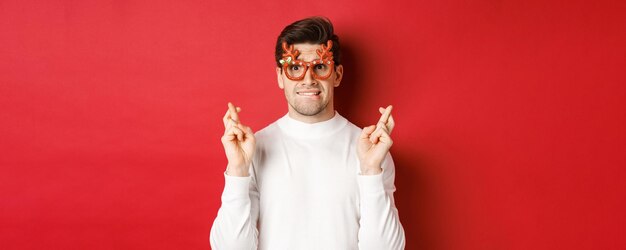 Closeup de um cara nervoso bonito em óculos de festa fazendo um desejo cruzando os dedos para boa sorte e lo