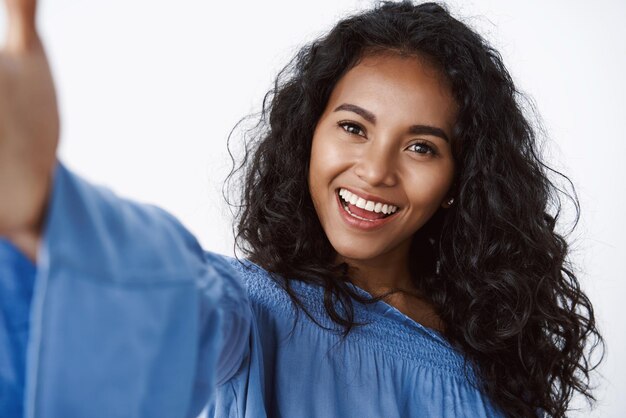 Closeup elegante e feminina afro-americana de cabelos encaracolados em blusa azul elegante estica a mão e segura o smartphone tomando selfie rindo e sorrindo fundo branco