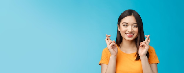 Closeup esperançosa linda garota asiática com cabelo escuro curto cruzar os dedos boa sorte orando sorrindo amplamente aguardando notícias positivas fielmente espero ganhar suporte fundo azul otimista animado