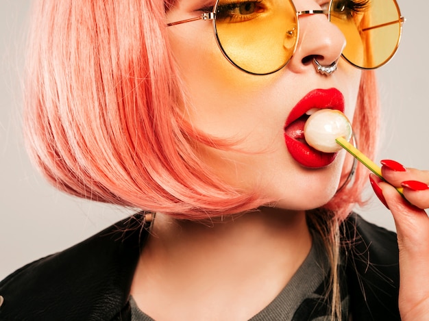 Closeup lábios da garota má jovem hippie bonita na jaqueta de couro preta na moda e brinco no nariz. Mulher sorridente despreocupada sexy posando no estúdio na peruca rosa. Modelo positivo lambendo doces redondos