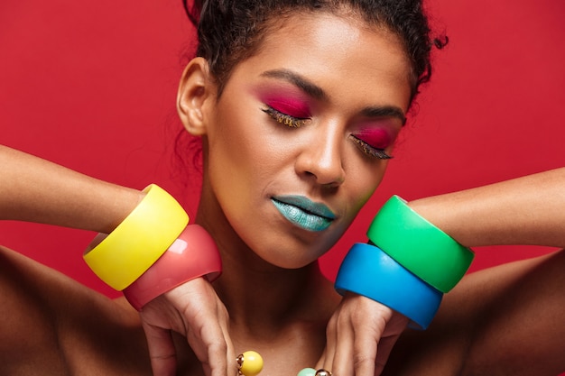 Foto grátis closeup mulata jovem sendo brilhante e elegante, gesticulando as mãos em pulseiras com os olhos fechados, muro vermelho