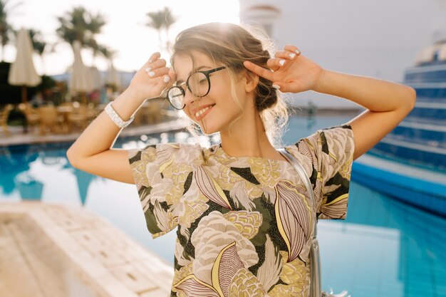 Closeup retrato de jovem bonito relaxante perto de bela piscina, resort. Senhora sorrindo, se divertindo, de férias, férias. Usando óculos da moda, camiseta, manicure curta branca.