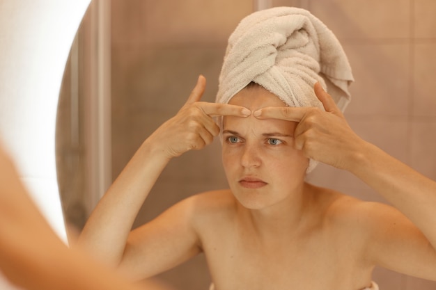 Foto grátis closeup retrato de jovem com acne na testa, olhando no espelho do banheiro com expressão preocupada, posando com ombros nus, procedimentos de beleza.